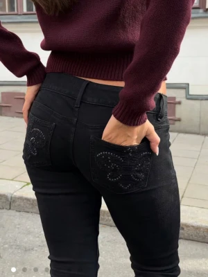 Gina tricot jeans - Säljer ett par svarta bootcut jeans med snygga broderade detaljer och små nitar på bakfickorna. Jeansen har låg midja och två knappar framtill. Materialet är stretchigt och bekvämt, perfekt för dig som gillar en cool och stilren look.