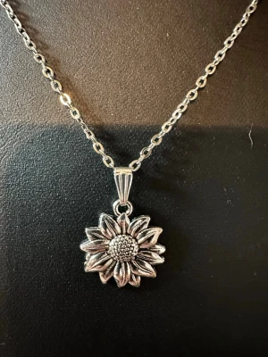 Silverfärgat halsband med solrosberlock - Stilrent halsband i silverfärg med en detaljrik solrosberlock. Kedjan har klassiska ovala länkar och berlocken är formad som en solros med tydliga kronblad och texturerad mitt. Perfekt accessoar för dig som gillar blommiga smycken och vill ha något unikt.