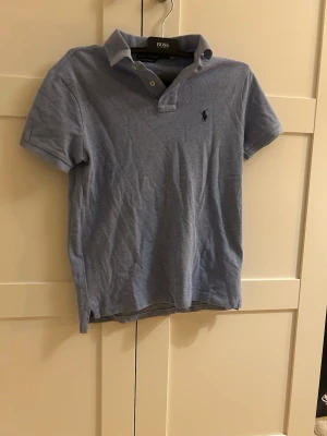 Ljusblå pikétröja från Ralph Lauren - Klassisk ljusblå pikétröja från Ralph Lauren med korta ärmar och den ikoniska broderade loggan på bröstet. Tröjan har krage och knappar vid halsen, perfekt för en avslappnad men ändå stilren look.