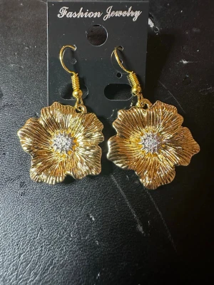 Guldiga blomörhängen  - Säljer ett par statement örhängen i form av stora blommor i guldfärgad metall med detaljer i silverfärg i mitten. Örhängena har krokfäste och en glansig finish som verkligen drar blickarna till sig. Perfekta för dig som vill ha något unikt och trendigt till din look.