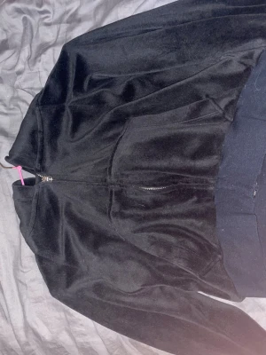 croppad Svart hoodie med strass-text - Säljer en croppad svart hoodie i mjukt velourmaterial med dragkedja framtill och fickor. På ryggen står det 'ANGELIC' i stora bokstäver av strass. Hoodien har en huva och bred mudd nedtill. Perfekt för dig som gillar streetwear med bling.