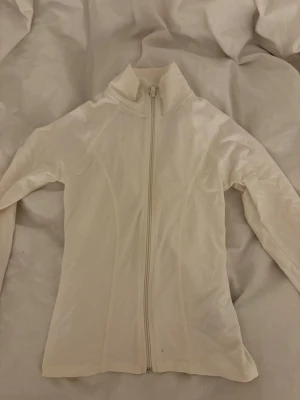 Vit zipjacka i syntetmaterial - Snygg vit jacka med hel dragkedja framtill och hög krage. Jackan har en figurnära passform och är tillverkad i ett mjukt syntetmaterial. Perfekt för lager på lager och enkel att matcha med olika outfits.