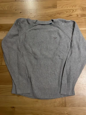 Ljusgrå stickad tröja från Gant - Ljusgrå stickad tröja från Gant med diskret logotyp på bröstet. Tröjan har ribbstickad struktur, rund halsringning och långa ärmar. Perfekt för lager-på-lager och enkel att matcha med jeans eller chinos.