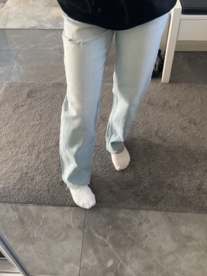 Ljusblå raka jeans  - Säljer ett par ljusblå jeans med rak passform och en liten slitning vid ena låret. Jeansen har klassisk femficksdesign, gylf med dragkedja och knapp samt bälteshällor. Perfekta för en avslappnad och trendig look. Från gina tricot i storlek 32