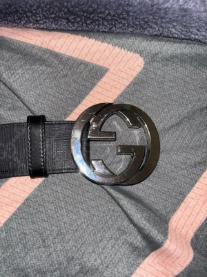 Svart Gucci bälte med GG-spänne - Snyggt svart bälte från Gucci med klassiskt GG-mönster längs hela remmen och ett stort, rundat metallspänne med GG-logga. Bältet är gjort i skinn och har en stilren, modern look som lyfter vilken outfit som helst.