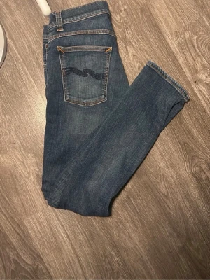 Blå jeans från Nudie Jeans - Säljer ett par klassiska blå jeans från Nudie Jeans med rak passform och snygga kontrastsömmar. Jeansen har en bakficka med Nudie Jeans karakteristiska brodyr och är gjorda i slitstarkt denimtyg. Perfekta för en avslappnad och stilren look.