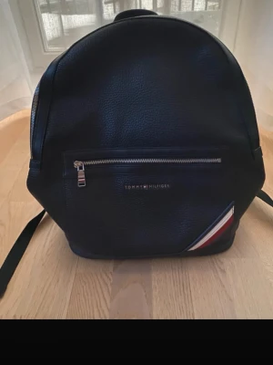 Svart ryggsäck Tommy Hilfiger - Snygg svart ryggsäck från Tommy Hilfiger i ett stilrent konstläder. Framtill finns en dragkedjeficka med diskret logga och klassiska Hilfiger-ränder i hörnet. Ryggsäcken har justerbara axelband och silverfärgade dragkedjor.