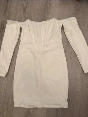 Vit offshoulder klänning med pärldetalj - Snygg vit kortklänning med offshoulder-design och långa ärmar. Klänningen har pärldetaljer längs kanten och markerad midja. Dragkedja baktill för enkel påklädning. Perfekt för dig som vill sticka ut med en elegant look.