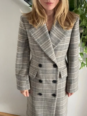 Grårutigt lång kappa med dubbelknäppning - Stilren lång kappa i grått och beige rutmönster med breda slag och dubbelknäppning framtill. Kappan har två fickor och markerad midja för en snygg siluett. Perfekt för dig som gillar klassisk men trendig stil.