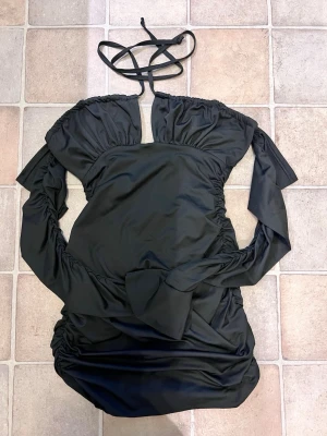 Svart omlottklänning med snörning - Cool svart kortklänning med omlott-design och snörning i halsen. Klänningen har långärmade ärmar med rynkade detaljer och en tight passform som framhäver siluetten. Materialet känns som polyester och ger en snygg glans. Perfekt för dig som vill sticka ut.