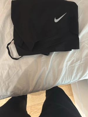 Svarta träningstights från Nike - Säljer ett par svarta träningstights från Nike med den klassiska vita swoosh-loggan på låret. Tightsen har en åtsittande passform och elastisk midja, perfekta för gymmet eller löpning. Materialet är stretchigt och andas bra.