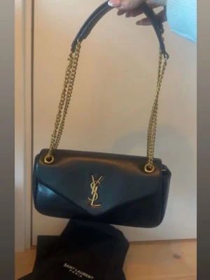 Svart YSL handväska i läder - Snygg svart handväska från Saint Laurent i mjukt läder med guldiga kedjor och ikoniska YSL-loggan framtill. Väskan har ett klassiskt kuvertlock och axelrem i kedja med läderdetalj. Perfekt för dig som gillar lyxiga accessoarer.