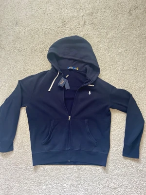Marinblå hoodie från Polo Ralph Lauren - Snygg marinblå hoodie från Polo Ralph Lauren med dragkedja framtill och vit logga broderad på bröstet. Hoodien har vita dragsnören i huvan och två fickor framtill. Tillverkad i mjukt bomullsmaterial för en skön och avslappnad look.
