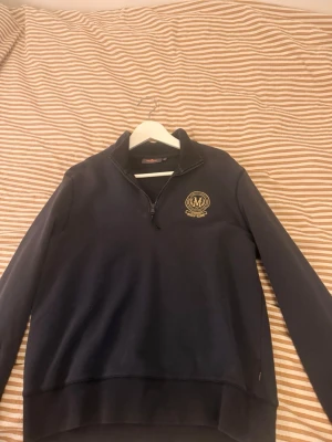Marinblå halvzip sweatshirt med brodyr - Snygg marinblå sweatshirt från Morris med halv dragkedja och guldfärgad broderad logga på bröstet. Tröjan har hög krage, långa ärmar och ribbade muddar. Perfekt för en avslappnad men stilren look. Använd fåtalet gånger