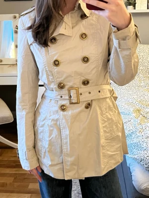 Beige trenchcoat med bälte - Stilren beige trenchcoat med dubbelknäppning och bälte i midjan. Klassisk krage, axelklaffar och dekorativa knappar i guldton. Tillverkad i ett lätt tyg, perfekt för lager på lager. Två sidofickor och justerbara ärmslut för extra stilpoäng.