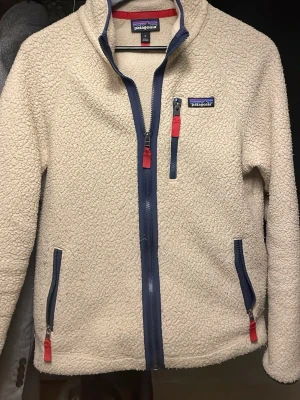 Vit fleecejacka från Patagonia - Mysig vit teddyjacka från Patagonia med blå detaljer och röda dragkedjor. Jackan har hög krage, tre fickor med dragkedja och en klassisk logga på bröstet. Perfekt för kyliga dagar och enkel att matcha med olika outfits.