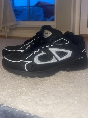 Svarta b30 sneakers med vita detaljer - Snygga svarta b30 med vita och grå detaljer samt stora CD-loggor på sidan. Skorna har en sportig siluett, meshpaneler och reflekterande inslag. Perfekta för dig som vill ha en exklusiv och trendig look. Skorna har använts 3 gånger. Skorna kan vara storleken 43-44 då jag inte är så säker. 