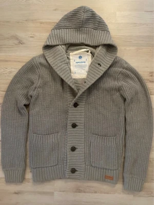 Grå stickad cardigan - Fet grå stickad hoodie med stora knappar framtill och två fickor. Tröjan har en bred ribbad kant vid ärmslut och nederkant samt en mysig huva. Perfekt nu för våren 🌱🌼🌳☀️