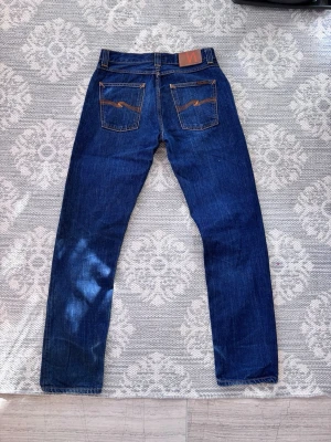 Mörkblå jeans från Nudie Jeans - Snygga mörkblå jeans från Nudie Jeans med klassisk femficksdesign och kontrastsömmar på bakfickorna. Jeansen har rak passform och är tillverkade i denim av bomull. Perfekta för en avslappnad och stilren look.