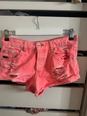 Korallfärgade jeans shorts med slitningar  - Coola rosa jeansshorts från pull and bear med slitna detaljer och fransig kant. Shortsen har klassisk femficksdesign, knapp och dragkedja framtill samt låg midja. Perfekta för sommardagar när du vill sticka ut lite extra.