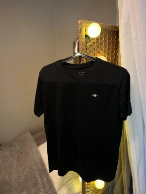 Svart t-shirt från Gant - Snygg svart t-shirt från Gant med liten broderad logga på bröstet. Klassisk rund halsringning och korta ärmar. Tillverkad i mjuk bomull som känns skön mot huden. Perfekt basplagg för en enkel och stilren look.