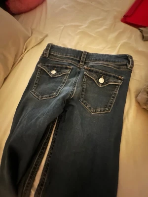 Mörkblå bootcut jeans med fickdetaljer - Snygga mörkblå bootcut jeans med dekorativa bakfickor med lock och silverfärgade knappar. De är lite slitna på slutet av byxorna på ena sidan av jeansen. 