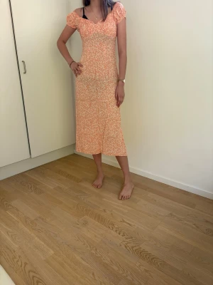 Blommig orange midiklänning - Söt blommig klänning i orange och vitt med korta puffärmar och smock upptill. Klänningen har en figurnära passform upptill och faller rakt ner till vaden. Perfekt för sommarens alla tillfällen!