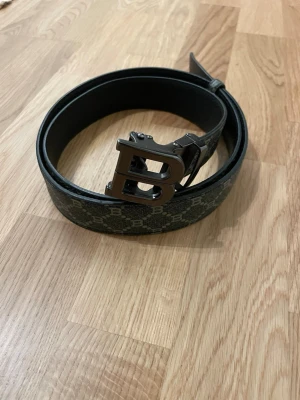 Svart skärp med B-spänne från Balmain - Snyggt svart skärp från Balmain med ett stort metallspänne i form av bokstaven B. Skärpet har ett diskret mönster med små B:n över hela bandet och är gjort i skinn. Perfekt accessoar för att lyfta din outfit.