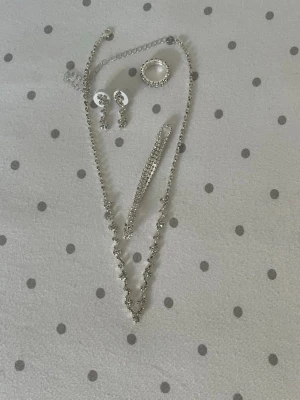 Glittrande smyckesset i silverfärg - Snyggt smyckesset med halsband, armband, ring och örhängen i silverfärg med gnistrande stenar. Perfekt för dig som gillar att sticka ut med bling och vill ha en matchande look. Klassisk design med rundade former och mycket glitter.