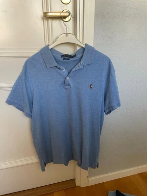 Ljusblå pikétröja från Polo Ralph Lauren - Klassisk ljusblå pikétröja från Polo Ralph Lauren med korta ärmar och krage. Tröjan har två knappar vid halsen och den ikoniska broderade loggan på bröstet. Perfekt till jeans eller shorts för en avslappnad men stilren look.