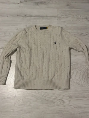 Vit kabelstickad tröja från Ralph Lauren - Kabelstickad tröja i offwhite från Ralph Lauren med rund halsringning och klassisk broderad logga på bröstet. Tröjan har ribbade muddar vid ärmslut och nederkant. Perfekt för dig som gillar en clean och tidlös stil.