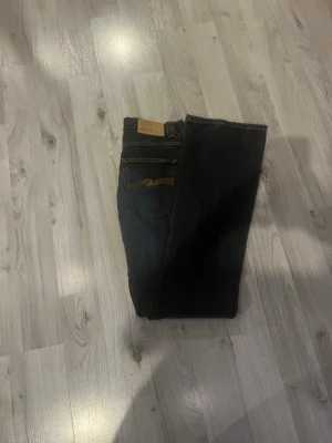 Svarta jeans från Nudie Jeans vintage  - Säljer ett par svarta jeans från Nudie Jeans med klassisk femficksmodell och gul kontrastsöm på bakfickan. Jeansen har rak passform och är tillverkade i denim. Perfekta för dig som gillar stilrena och tidlösa plagg.