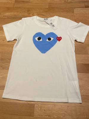 Vit Comme des Garçons Play t-shirt - Säljer en vit t-shirt från Comme des Garçons Play med stort blått hjärta med ögon och ett litet rött hjärta på bröstet. Klassisk rund hals och korta ärmar. Perfekt statement-plagg för dig som gillar streetwear och coola prints.