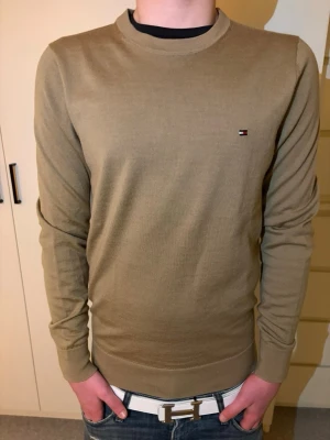 Beige tröja från Tommy Hilfiger - Snygg beige tröja från Tommy Hilfiger med rund halsringning och diskret logga på bröstet. Tröjan har långa ärmar och ribbade muddar vid ärmslut och nederkant. Perfekt för en clean och stilren look.