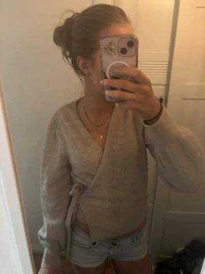 Beige omlottkofta från &other stories - Säljer en beige stickad omlottkofta från  &other stories med knytband i midjan och långa ärmar. Koftan har en djup v-ringning och ribbade muddar. Perfekt att slänga över en topp för en chill look.