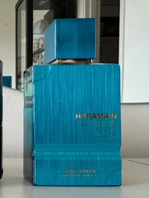 Haramain Amber Oud Aqua Dubai parfym - Den är använd fåtal gånger och du har minst 70 mL En lyxig parfymflaska från Al Haramain, Amber Oud Aqua Dubai. Flaskan är rektangulär med en metallisk blå finish och matchande blått lock. Texten är i guld och flaskan har en modern, stilren design i glas och metall.