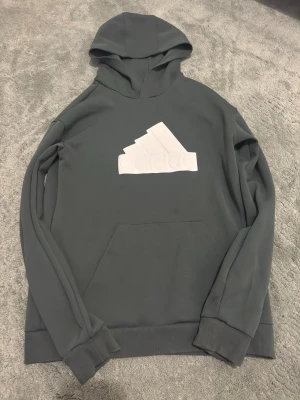 Adidas hoodie - Snygg mörkgrå hoodie från Adidas med stor vit logga på bröstet. Klassisk känguruficka framtill och ribbade muddar vid ärmar och nederkant. Tillverkad i mjukt material som känns skönt mot huden. Perfekt för en avslappnad och sportig stil.