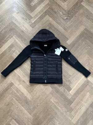 Svart pufferjacka med huva från Moncler - Säljer en svart pufferjacka från Moncler med huva och quiltad front. Jackan har stickade ärmar med Moncler-logga på ena armen och en praktisk ficka med dragkedja. Perfekt för höst/vår dagar eller lite kyligare sommardagar 😁