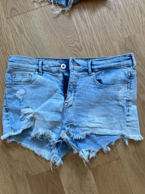 Ljusa denimshorts med fransar från H&M - Säljer ett par ljusa blå denimshorts från H&M med rå franskant och slitna detaljer. Shortsen har klassisk femficksdesign, knapp och dragkedja framtill samt låg midja. Perfekta för sommarens alla äventyr!