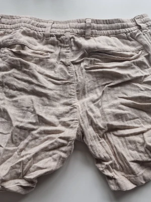 Beige linneshorts med snörning - Snygga beige shorts i linne med elastisk midja och snörning framtill. Shortsen har två bakfickor med lock och en avslappnad passform, perfekta för varma dagar.