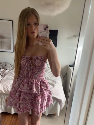 Blommig volangklänning med snörning - Säljer nu denna fantastiska rosablommiga miniklänning. Modellen är axelbandslös med ett stretchigt material med fina volanger på kjolen som ger en jättesöt siluett.Helt perfekt för sommarens middagar på stranden eller semesterresor! 
