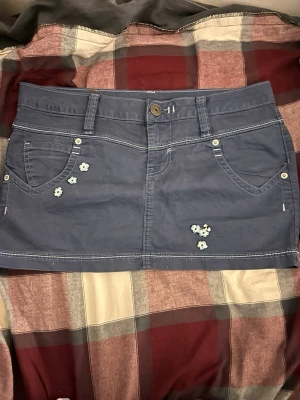 Blå kortkjol med blommor från Replay - Snygg blå kortkjol från Replay med små broderade blommor framtill. Kjolen har klassisk femficksdesign, bälteshällor och kontrastsömmar. Perfekt för dig som gillar en avslappnad men trendig stil.