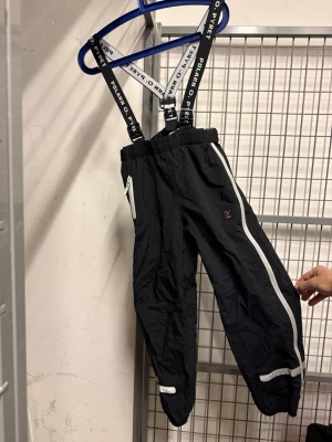 Svarta skalbyxor med hängslen Polarn O. Pyret - Svarta skalbyxor från Polarn O. Pyret med vita detaljer och reflexer. Justerbara svarta hängslen med logga, resår i midjan och dragkedja i sidan. Hög kvalitrt 