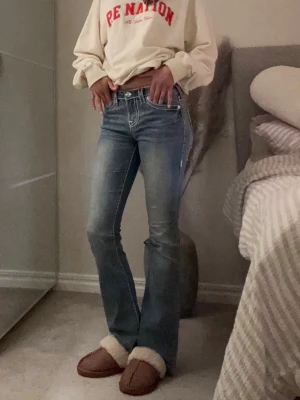 True Religion bootcut jeans blå - Säljer ett par blå bootcut jeans från True Religion med ikoniska röda och vita kontrastsömmar på bakfickorna. Jeansen har låg midja och klassisk femficksdesign. Perfekta för dig som gillar en avslappnad men trendig look. 