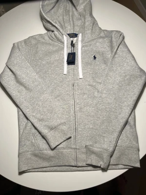 Grå hoodie från Polo Ralph Lauren - Snygg ljusgrå hoodie från Polo Ralph Lauren med dragkedja, vit dragsko och klassisk blå broderad logga på bröstet. Mjuk bomullsblandning, huva och fickor framtill. Perfekt för en chill och stilren look.