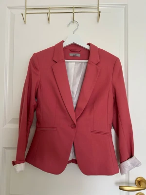 Rosa kavaj med klassisk passform - Snygg rosa kavaj med figursydd passform och klassisk krage. Kavajen har en knapp framtill, två fickor och ljusrosa foder. Perfekt för att lyfta din outfit med lite färg.
