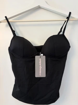 Svart top från Missguided - Snygg svart top från Missguided med tunna axelband och vadderade kupor. Tillverkad i mjukt stretchigt material som sitter bekvämt mot kroppen. Metallogo bak för extra detalj. Perfekt för dig som vill ha en stilren och enkel look.