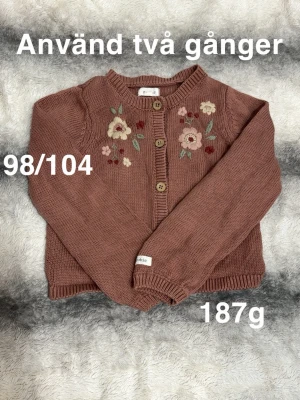 Brun kofta med blommor från Newbie - Söt brun kofta från Newbie med broderade blommor framtill. Koftan har rund hals, ribbade muddar och knappar i trä-look. Perfekt för lager-på-lager och ger en gullig touch till outfiten.🌸 Använd två gånger. Storlek 98/104
