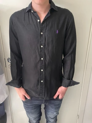 Svart linneskjorta från Polo Ralph Lauren - Svart skjorta i linne från Polo Ralph Lauren med slim fit-passform. Klassisk krage, vita knappar och lila broderad logga på bröstet. Perfekt för en stilren och avslappnad look. Modellen på bilden är 185 cm och väger 75 kg.
