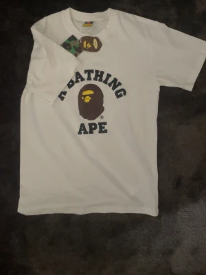 Vit BAPE t-shirt med logga - Snygg vit t-shirt från A Bathing Ape med den klassiska BAPE-loggan och apans ansikte tryckt på bröstet. T-shirten har rund hals och korta ärmar. Perfekt för dig som gillar streetwear och vill sticka ut med en ikonisk design.